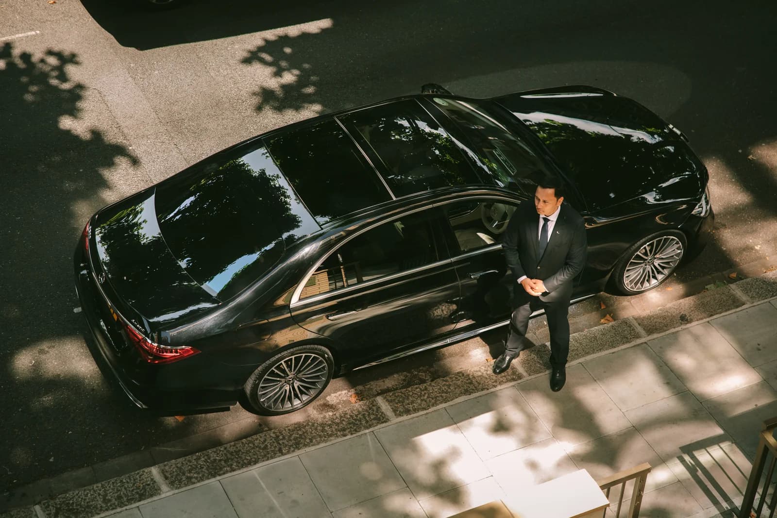 Professional chauffeur standing by a black Mercedes sedan — top-down aerial view